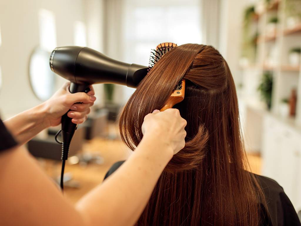 One stylist blow-drying a client's hair with a round wooden brush, seen from behind
